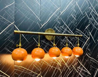 May include: A gold metal light fixture with five orange glass globes. The fixture is mounted on a black wall with a white and gold pattern.