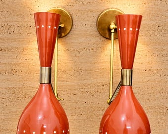 Op de afbeelding: Een paar wandlampen met oranje dubbele kegelvormige kappen met witte stippen. Elke lamp wordt ondersteund door een messingkleurige metalen arm en een ronde muurplaat en verspreidt een warm licht via de openingen aan de uiteinden.