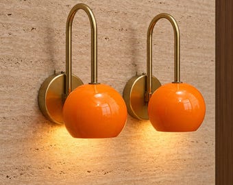 May include: A pair of wall sconces featuring a brass-toned circular mounting plate and a curved arm that supports a glossy, spherical orange glass shade. Each lamp emits a warm, downward-facing light, highlighting the smooth, reflective surface of the orange globes.