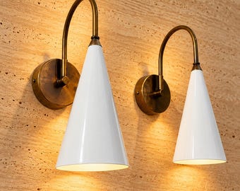May include: Two wall sconces featuring white conical shades and curved antique brass arms. Each lamp is mounted on a circular brass base, emitting a warm downward glow from the open bottom of the cone-shaped fixture.