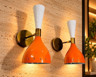 May include: Two wall-mounted lamps featuring bright orange, bell-shaped shades with small perforations along the bottom edge, connected to white, conical top sections by brass-coloured hardware and circular wall plates.
