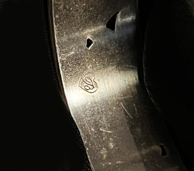 Pu&ograve; includere: Primo piano di una fascia di metallo argentata con un marchio a forma di cuore che contiene la lettera "S".