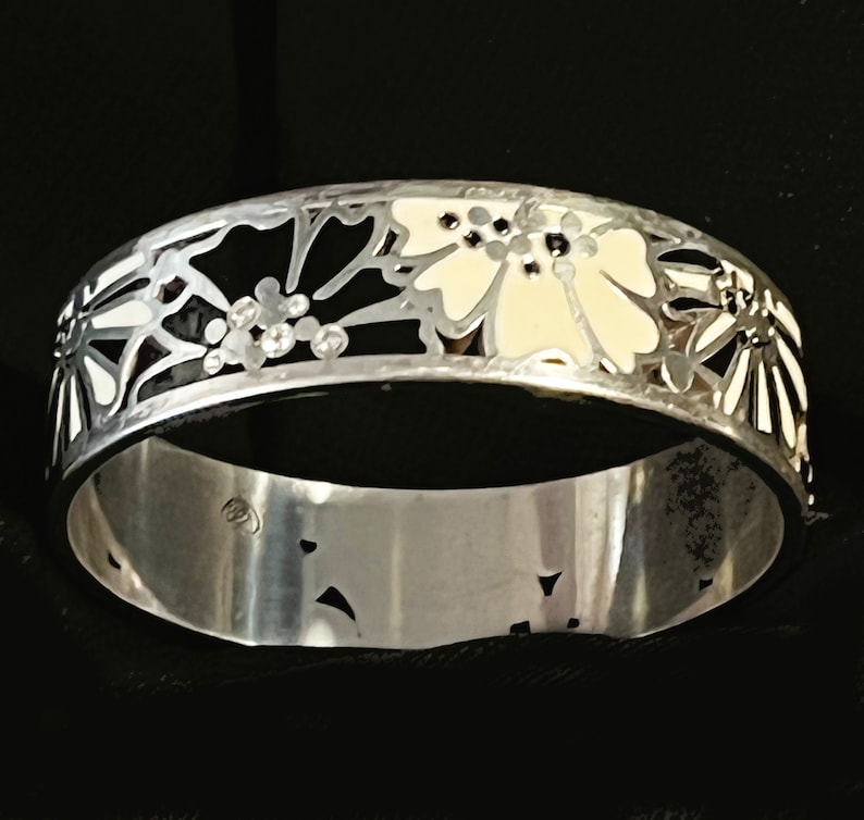 Pu&ograve; includere: Un braccialetto rigido in argento con un design floreale bianco e nero. I fiori sono contornati da argento e riempiti di smalto bianco.