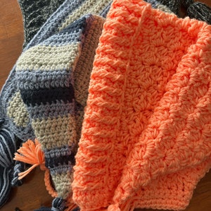 May include: Three crocheted hats with tassels. One hat is a striped pattern in shades of grey and blue, another is a solid peach colour, and the third is a dark grey with a textured pattern.