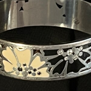 Pu&ograve; includere: Un braccialetto rigido in argento con disegni floreali in smalto nero e bianco. Il braccialetto ha un design delicato e intricato con un motivo floreale.