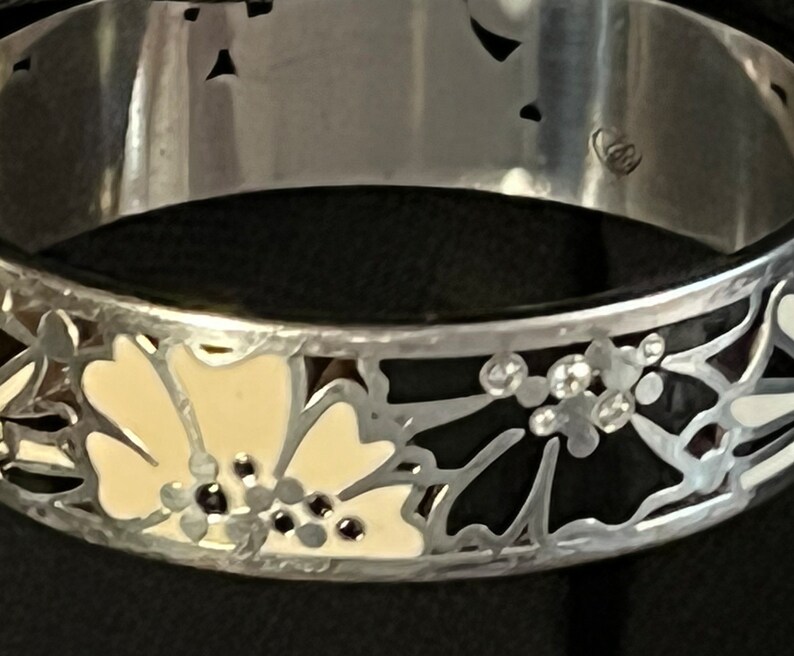 Pu&ograve; includere: Un braccialetto rigido in argento con disegni floreali in smalto nero e bianco. Il braccialetto ha un design delicato e intricato con un motivo floreale.