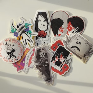 Könnte beinhalten: Ein Set aus zehn bunten Aufklebern mit verschiedenen Illustrationen und Texten. Die Aufkleber zeigen einen Schädel, ein Herz, einen Schmetterling, einen Sarg und ein Paar. Der Text auf den Aufklebern lautet "Cause I'm Still Here", "Our Lady Of Sorrows", "Am Not Afraid", "Awake", "The Lovers" und "Look Alive, Sunshine".