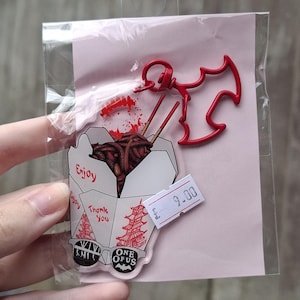 May include: A clear acrylic pin featuring a cartoon takeout container with noodles and chopsticks. The pin includes the text "Enjoy" and "Thank You". A red bat-shaped charm and a price tag are also included.