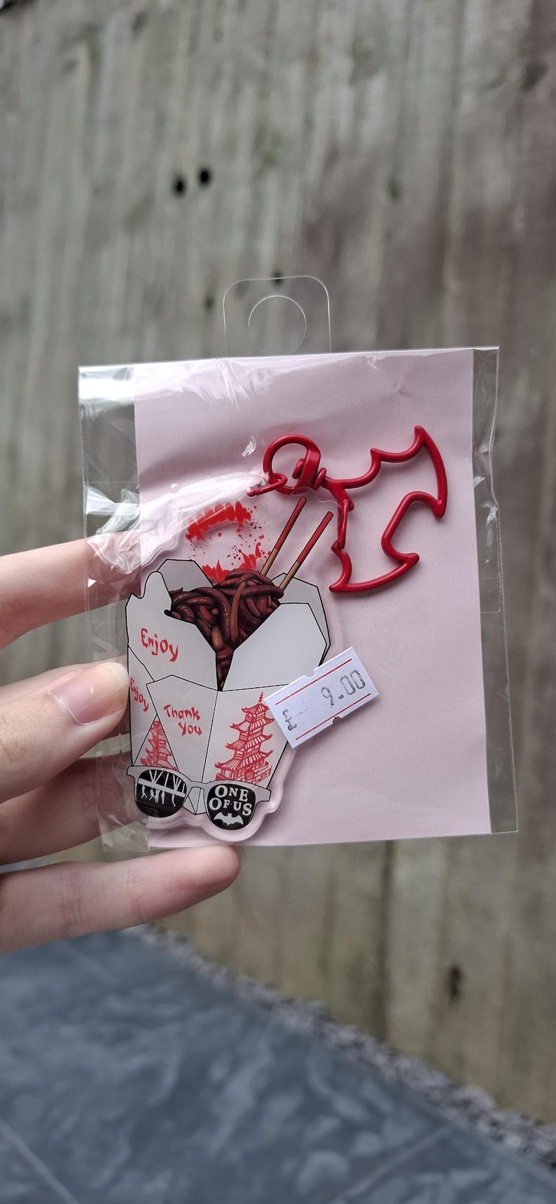 May include: A clear acrylic pin featuring a cartoon takeout container with noodles and chopsticks. The pin includes the text "Enjoy" and "Thank You". A red bat-shaped charm and a price tag are also included.
