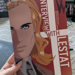 Könnte beinhalten: Ein Buchcover mit dem Titel Interview with Lestat, das eine stilisierte Illustration einer Person mit langem blondem Haar und blauen Augen zeigt. Das Design umfasst rote und weiße geometrische Blöcke mit dem Text geschrieben von Daniel Molloy und annotiert von Lestat de Lioncourt.
