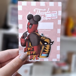 May include: A pink and white checkered card with the text "Thank You" and a price tag that says "3.50". The card features a cartoon illustration of Deadpool wearing Mickey Mouse ears and a backpack. The text "Bye Bye Bye" is printed on a sticker below the illustration.