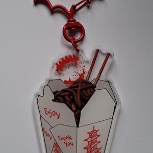 May include: A red Batman-shaped keychain with a clear acrylic charm. The charm features a takeout box graphic with noodles, chopsticks, and the words "Enjoy" and "Thank You". The bottom has the text "ONE OF US".