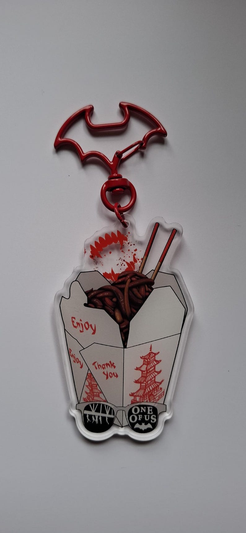 May include: A red Batman-shaped keychain with a clear acrylic charm. The charm features a takeout box graphic with noodles, chopsticks, and the words "Enjoy" and "Thank You". The bottom has the text "ONE OF US".