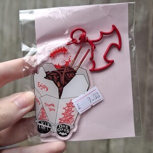 May include: A clear plastic package containing a red bat-shaped charm and a food-themed acrylic pin. The pin features a takeout box with noodles, chopsticks, and the words "Enjoy" and "Thank You".