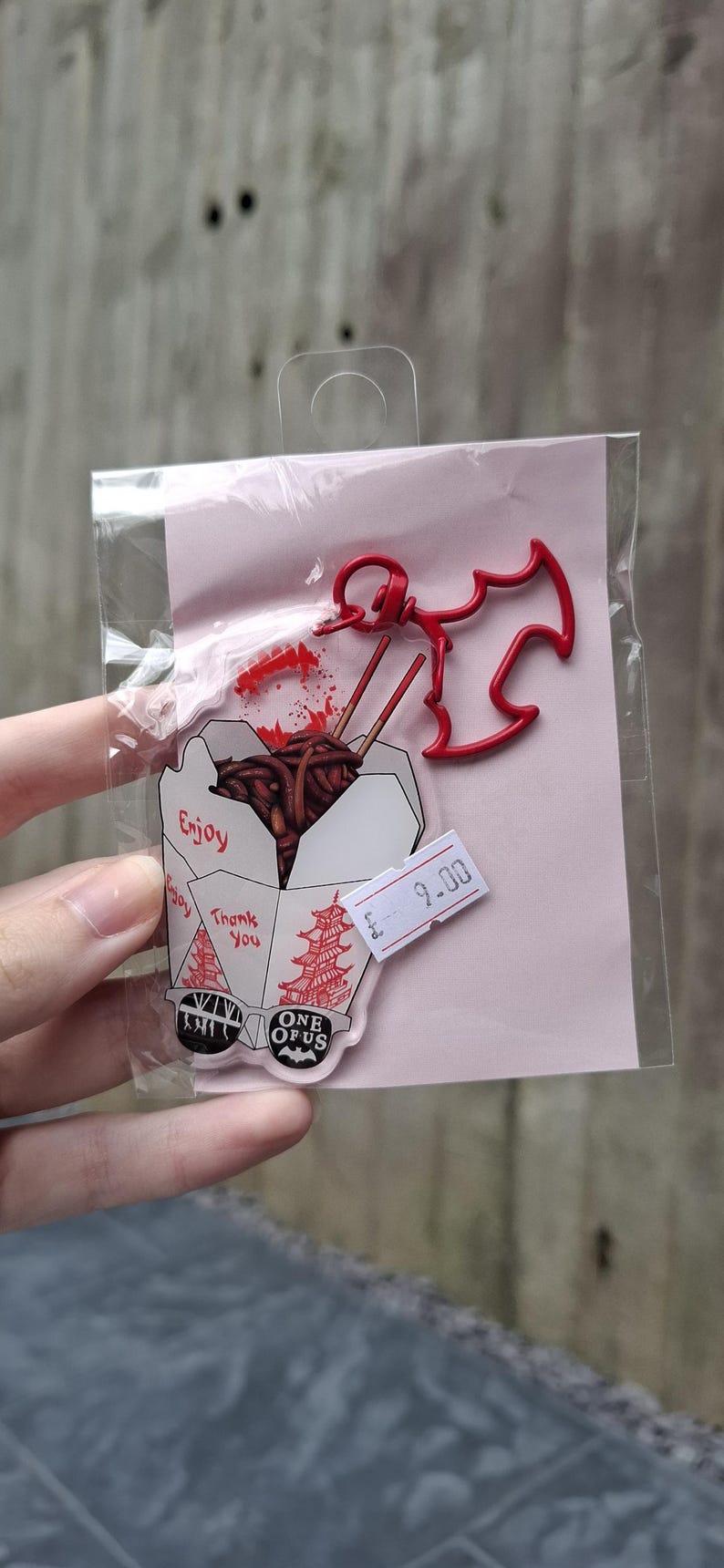 May include: A clear plastic package containing a red bat-shaped charm and a food-themed acrylic pin. The pin features a takeout box with noodles, chopsticks, and the words "Enjoy" and "Thank You".