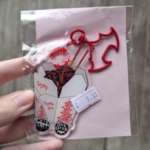 May include: A clear acrylic charm featuring a takeout box with noodles and chopsticks, a red bat-shaped charm, and the text "Enjoy" and "Thank You". The charm is packaged on a pink backing.