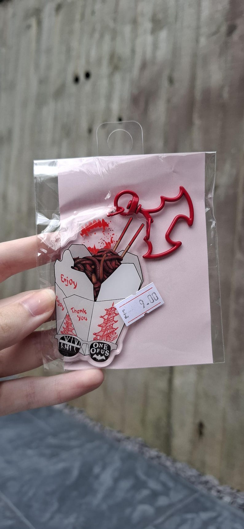 May include: A clear acrylic charm featuring a takeout box with noodles and chopsticks, a red bat-shaped charm, and the text "Enjoy" and "Thank You". The charm is packaged on a pink backing.