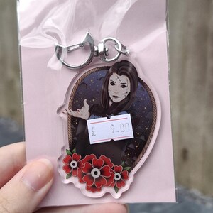 May include: A clear acrylic keychain with a woman's portrait, featuring cracked face makeup and dark hair. The keychain has a silver clasp and a floral design. The price tag shows "9.00".