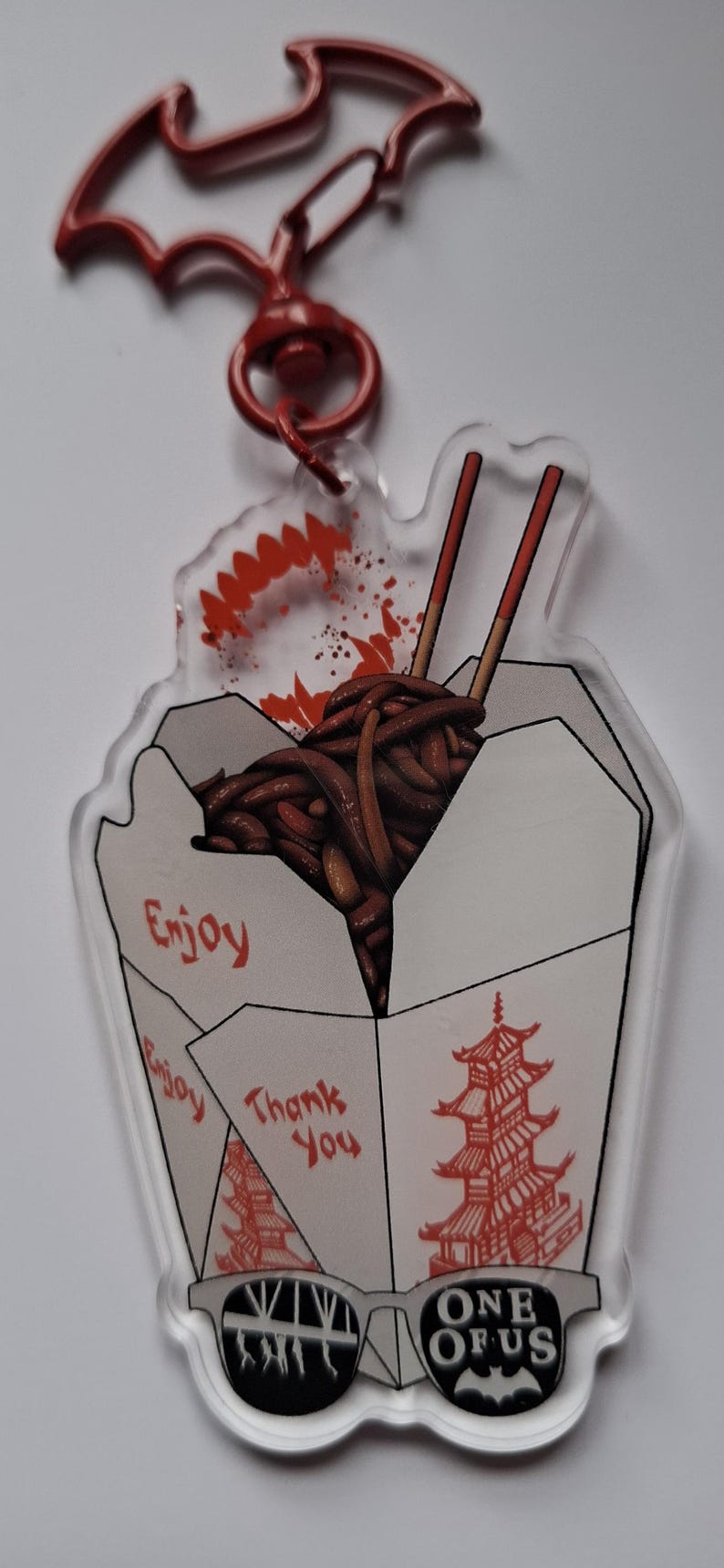 May include: Clear acrylic keychain featuring a Chinese takeout box filled with noodles and chopsticks. The keychain has a red bat-shaped clasp and includes the text "Enjoy", "Thank You", and "One of Us".