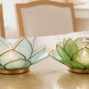 May include: Two lotus flower candle holders made of translucent light green and dark green capiz shell petals with dark metal edging and a circular gold base. Each holder features a central light source that illuminates the delicate shell petals.