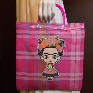 May include: Pink and white plaid tote bag with a clear plastic front and pink handles. The bag features an embroidered patch of a cartoon Frida Kahlo with flowers in her hair.