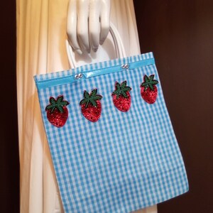 May include: A blue and white gingham tote bag with a blue ribbon and four red strawberry appliques.