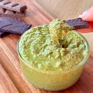 May include: A clear glass bowl filled with green dip, with a piece of dark chocolate being dipped into it. Two types of chocolate bars are on a wooden tray. The dip has a thick, textured consistency.