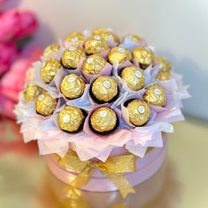 Puede incluir: Una caja de regalo rosa llena de un ramo de bombones de chocolate dorados envueltos en papel de seda blanco. La caja est&aacute; atada con una cinta dorada.