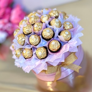 Puede incluir: Una caja redonda rosa llena de un ramo de bolas de chocolate doradas envueltas en papel de seda blanco y morado. La caja tiene una cinta dorada alrededor.