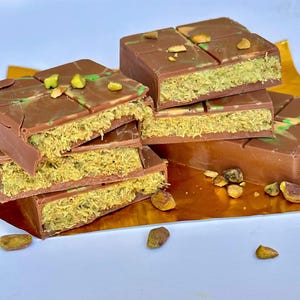 May include: A stack of chocolate bars with pistachio filling. The bars are broken in half, revealing the green pistachio filling. The bars are on a gold foil surface with pistachio nuts scattered around.