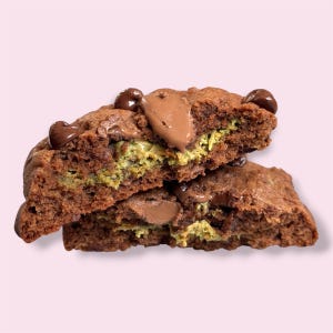 May include: Two halves of a chocolate cookie with a green filling, stacked on top of each other. The cookie has chocolate chips and a melted chocolate topping. The background is a solid light pink.