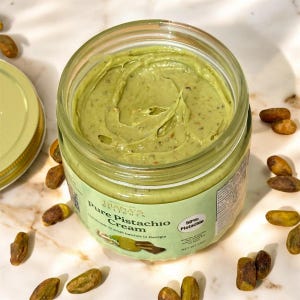 Puede incluir: Un tarro de cristal abierto con crema de pistacho verde, con trozos de nuez visibles. La etiqueta dice "Pure Pistachio Cream" y "50% Pistachio". Pistachos dispersos rodean el tarro sobre una superficie de mármol. La tapa del tarro es visible a la izquierda.