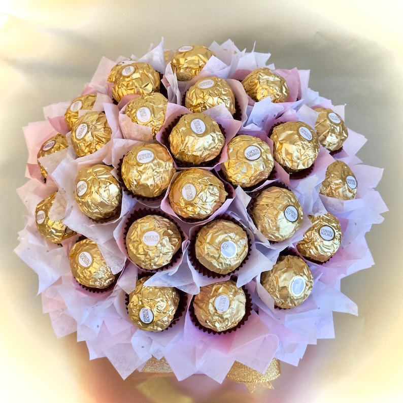 Puede incluir: Un ramo de bombones de chocolate dorados envueltos en papel de aluminio, dispuestos en un ramo de papel de seda rosa y blanco.