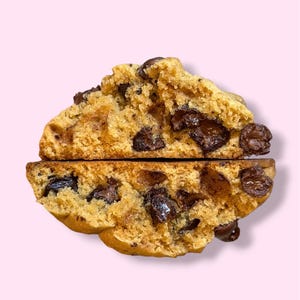 May include: A chocolate chip cookie, cut in half to show the inside texture. The cookie is a light brown colour with dark chocolate chips. The background is a pale pink.