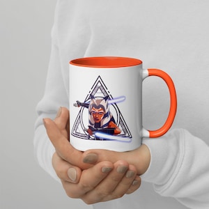 May include: A white ceramic mug with an orange rim and handle. The mug features a black and white illustration of a female character with orange skin and blue eyes, holding two lightsabers. The character is surrounded by a black triangle with a white outline.