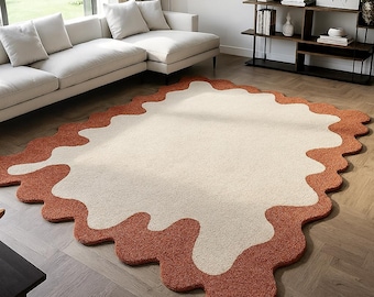May include: A large, abstract rug with a cream-colored center and a rust-colored, wavy border. The rug is placed on a hardwood floor in a living room setting, with a white sofa and shelving unit in the background. The rug's design adds a modern touch to the room.