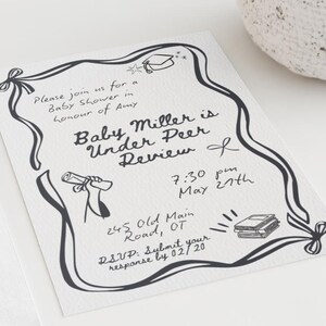 Può includere: Un invito bianco con testo e illustrazioni scritti a mano in nero per un baby shower. L'invito è accanto a una busta bianca e un vaso bianco strutturato. Il testo recita "Baby Miller is Under Peer Review".