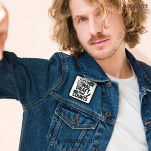 May include: A denim jacket with a patch that reads "IT'S THE FINAL DRAFT THAT COUNTS" in black and green. The patch is square and has a checklist design. The jacket has metal buttons and a classic collar.