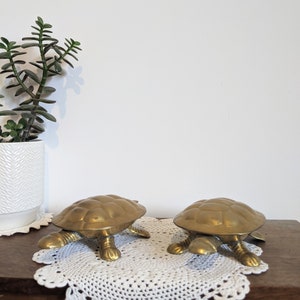 May include: Two gold-toned metal turtle figurines on a white crocheted doily. The turtles are facing the same direction and have detailed shell patterns.