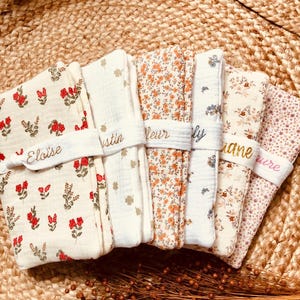 Puede incluir: Una pila de cinco mantas para beb&eacute; con estampados florales, cada una asegurada con una banda blanca bordada con un nombre. Las mantas presentan varios dise&ntilde;os florales en rojo, naranja y rosa sobre un fondo crema. Sobre una superficie tejida.