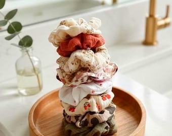 May include: A stack of seven colorful scrunchies on a wooden tray. The scrunchies are in various colors and patterns, including floral, polka dot, and solid colors. The tray is sitting on a white countertop in a bathroom setting.