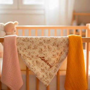 Peut inclure: Trois langes en mousseline posés sur un lit en bois. Un lange est rose pâle uni, un autre est jaune moutarde uni, et celui du centre présente un motif floral marron avec le prénom "Lodane" brodé en lettres cursives marron.