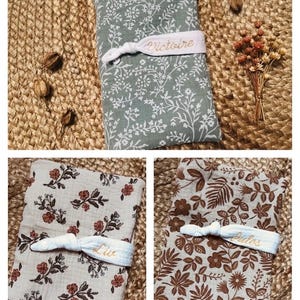 Puede incluir: Tres mantas para beb&eacute; con estampados florales en verde salvia, beige y marr&oacute;n, cada una asegurada con una banda el&aacute;stica blanca. Las bandas est&aacute;n bordadas con nombres. Las mantas se exhiben sobre una superficie de yute tejido.
