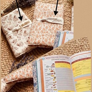 Puede incluir: Se muestran dos cubiertas de libros de tela floral con los nombres bordados "Louane" y "Fleur". Las cubiertas son en tonos crema y melocot&oacute;n, con una cinta de tela a juego. Se ve un libro abierto con p&aacute;ginas coloridas, junto con lavanda seca.