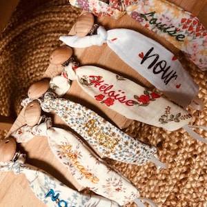 May include: A collection of six fabric pacifier clips featuring wooden circular clips and various floral or patterned cotton straps. Each strap is embroidered with a unique name including Louise, Nour, Héloïse, and Noa in different thread colors.