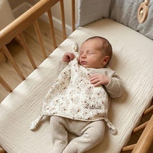 Peut inclure: Une grenouillère à manches longues en tricot beige avec pieds et une couverture de sécurité carrée en tissu à motif floral dans des tons marron et beige. La couverture a des coins noués et le prénom Arthur brodé en écriture marron sur le devant.