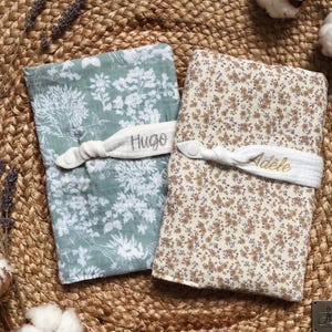 Puede incluir: Dos mantas para beb&eacute;, una en verde salvia con un estampado floral blanco y la otra en crema con un estampado floral marr&oacute;n. Cada manta est&aacute; asegurada con una banda de tela blanca bordada con un nombre. La manta con el estampado verde salvia tiene el nombre "Hugo". La otra manta tiene el nombre "Ad&egrave;le".