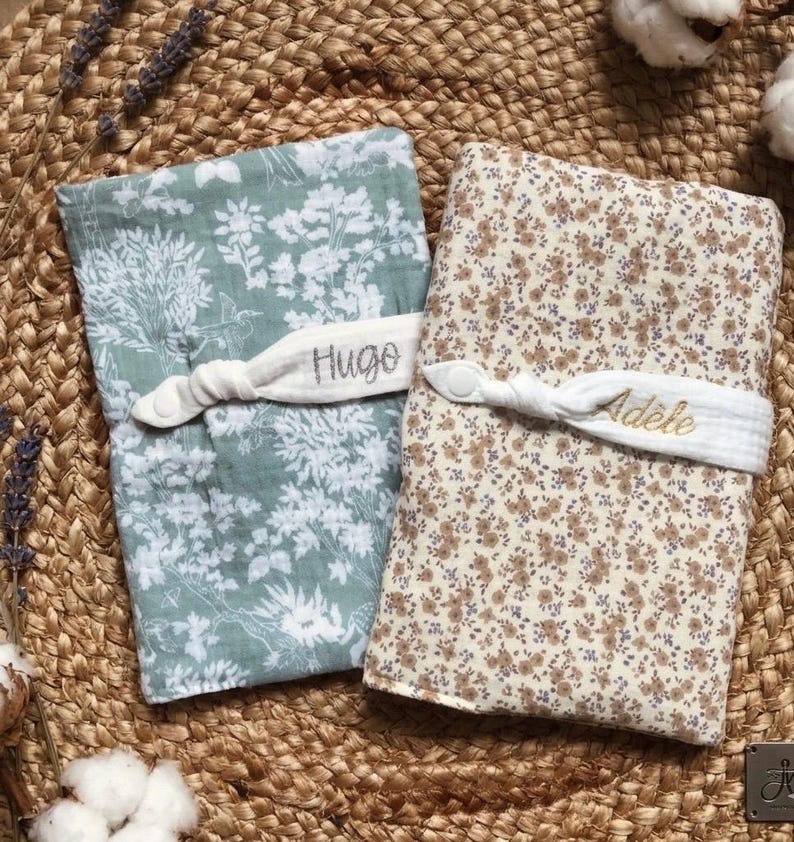 Puede incluir: Dos mantas para beb&eacute;, una en verde salvia con un estampado floral blanco y la otra en crema con un estampado floral marr&oacute;n. Cada manta est&aacute; asegurada con una banda de tela blanca bordada con un nombre. La manta con el estampado verde salvia tiene el nombre "Hugo". La otra manta tiene el nombre "Ad&egrave;le".