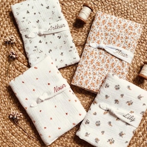 Puede incluir: Cuatro mantas de beb&eacute; plegadas con estampados florales y de coraz&oacute;n. Cada manta est&aacute; atada con una banda blanca bordada con un nombre: Arthur, Fleur, Nour y Lily. Las mantas son blancas, naranjas y marrones.