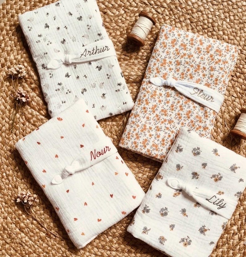 Puede incluir: Cuatro mantas de beb&eacute; plegadas con estampados florales y de coraz&oacute;n. Cada manta est&aacute; atada con una banda blanca bordada con un nombre: Arthur, Fleur, Nour y Lily. Las mantas son blancas, naranjas y marrones.